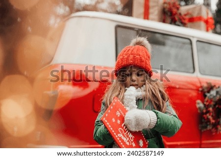 Female child celebrating Christmas and New Year winter holidays season outdoor. Little girl joyful spending time on open air holding letter to Santa in red envelope and waiting for miracle