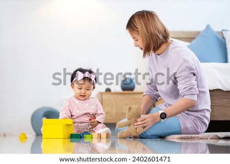 Asian mother is playing with her pretty smiling baby daughter with wooden toy block while spending quality time in bed for family happiness and parenting