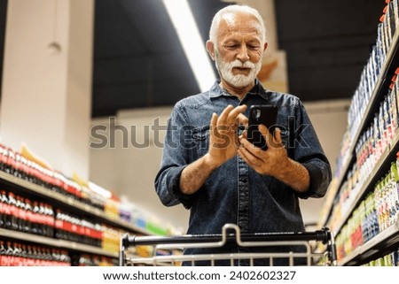 Senior Man doing grocery shopping at the supermarket, he is using apps on his smartphone. Male customer shopping with smartphone checklist, taking products from shelf at the shop.