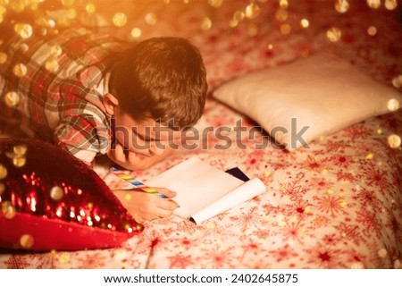 Close-up Cute little boy in red checkered pajamas writes a letter to Santa Claus while lying on the bed in the background of a flickering garland