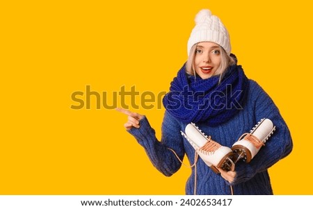 Beautiful young woman with ice skates pointing at something on yellow background