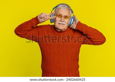 Excited senior stylish old man listening music via headphones and dancing disco party fooling around having fun expressive gesticulating hands. Happy mature grandfather pensioner on yellow background
