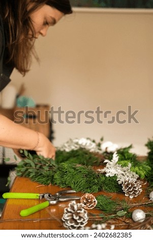 Vertical photo of a young woman in an apron assembling a Christmas wreath from fir branches. Decorations for the New Year are scattered on the table: pine cones, Christmas tree balls and a star.