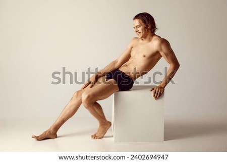 Young man with fit, muscular body sitting shirtless in underwear against grey studio background. Concept of men's beauty, health, body care, sportive lifestyle. Empty space to insert text Royalty-Free Stock Photo #2402694947