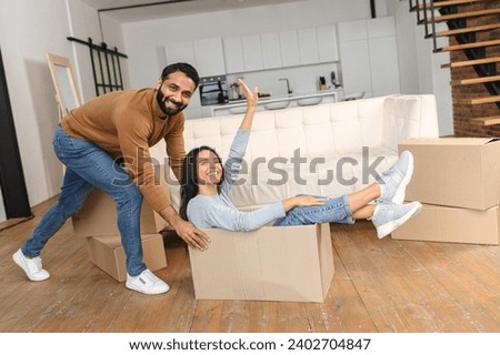 Happy Indian couple having fun while unpacking belongings on moving day. Excited wife riding in cardboard box while her husband push it in new house apartment Royalty-Free Stock Photo #2402704847