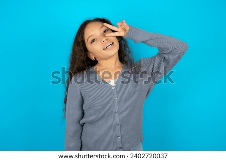 Beautiful kid girl wearing casual jacket over blue background Doing peace symbol with fingers over face, smiling cheerful showing victory