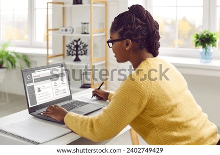 Young African American business woman sitting at working desk in office, using laptop computer, browsing online banking website, updating personal details and taking notes in notebook Royalty-Free Stock Photo #2402749429