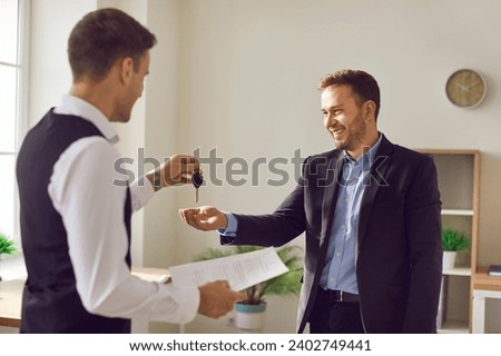 Portrait of professional man realtor agent standing in office with signed contract and giving house key to a happy smiling man customer and new homeowner. Real estate market or car purchase concept.
