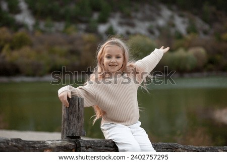 Cute blond girl, 7-years-old having fun on the pier of the pond. The kid dress on boots, delicate shades of clothes, sweater and jeans
