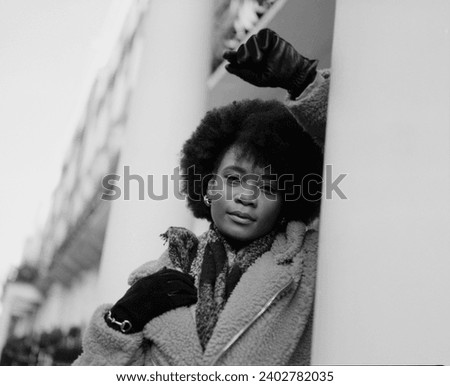 Black and white portrait of black woman leaning on Victorian building columns. It is a cold winter day and she is wearing coat, scarf and gloves. Portrait made with analog camera.