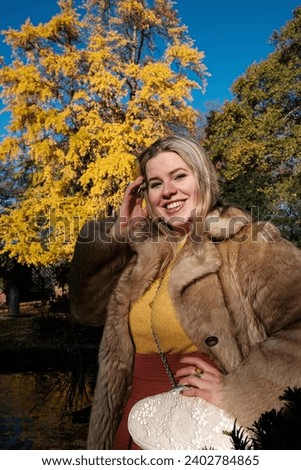 Smiling of friendly young woman posing in an autumn park in London. It is a cold morning day and she is wearing a faux fur coat and warm sweater. She is also wearing a shoulder bag.