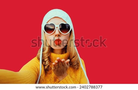Winter portrait of beautiful young woman stretching hand for taking selfie with mobile phone wearing yellow knitted sweater, white hat, heart shaped sunglasses on red studio background