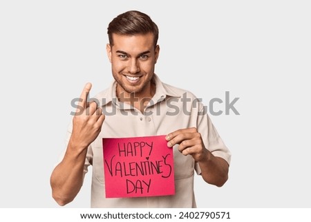 Young man with Valentine's sign pointing with finger at you as if inviting come closer.