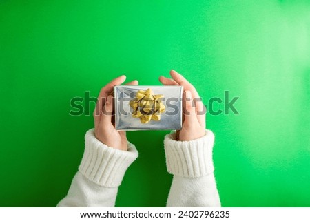 First person top view photo of hands holding silver giftbox with golden ribbon bow over shiny golden sequins on isolated green background
