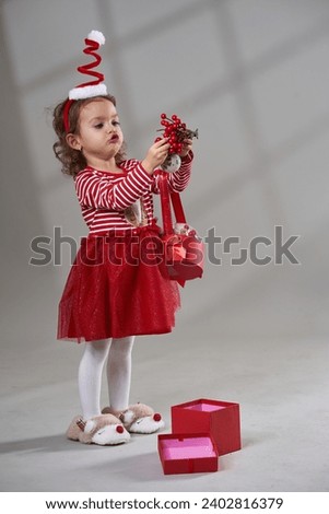 Portrait of an adorable blonde curly hair little girl in red dress and hat opening christmas present on gray background, studio shot