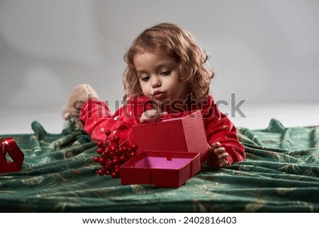 Portrait of an adorable blonde curly hair little girl in red dress and hat opening christmas present on gray background, studio shot