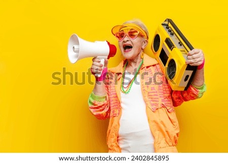 funny crazy old granny with tape recorder in sports hipster clothes listens to rock music and announces into megaphone on yellow background, elderly woman screams into loudspeaker at party