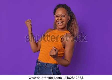 Young overjoyed beautiful African American woman student happily waves arms and experiences euphoria after receiving large educational grant to enter university stands on plain purple background.