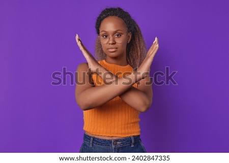 Young beautiful attractive African American woman demonstrates stop gesture by crossing arms and calling to cease racial or gender discrimination caused by lack of tolerance stands in purple studio. Royalty-Free Stock Photo #2402847335