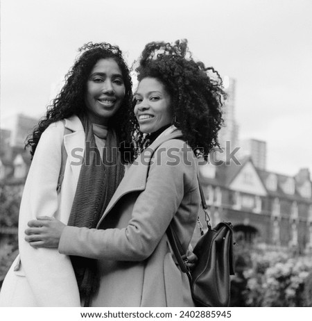 Analog portrait of two female friends posing in the street with affection. They are hugging each and looking at camera. It is a monochromatic image made with a medium format camera.