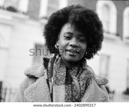 Analog portrait of smiling young curly black woman looking straight to camera in the street. She has a building behind and she is wearing winter outfit. Image made with medium format analog camera.