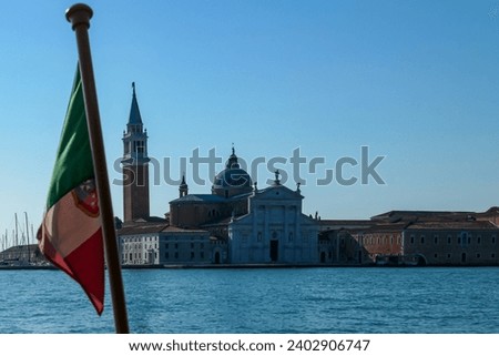 Waving Italian flag with scenic view church on the Island of San Giorgio Maggiore, Venice, Veneto, Northern Italy, Europe. Perspective from ferry boat trip. Patriotic banner moving to the wind
