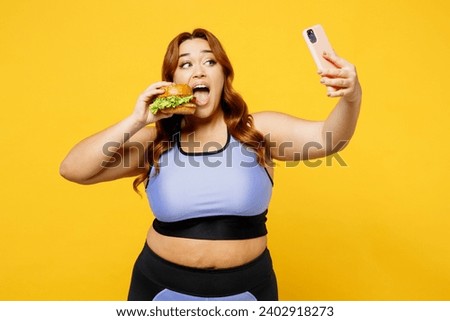 Young chubby overweight plus size big fat fit woman wear blue top warm up train eat burger do selfie shot on mobile cell phone isolated on plain yellow background studio home gym Workout sport concept