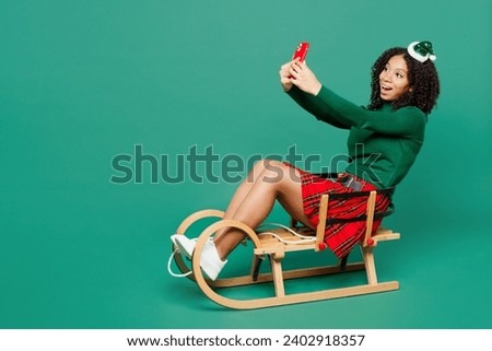 Full body merry little kid teen girl wears hat casual clothes posing sledding doing selfie shot on mobile cell phone isolated on plain green background studio. Happy New Year Christmas holiday concept