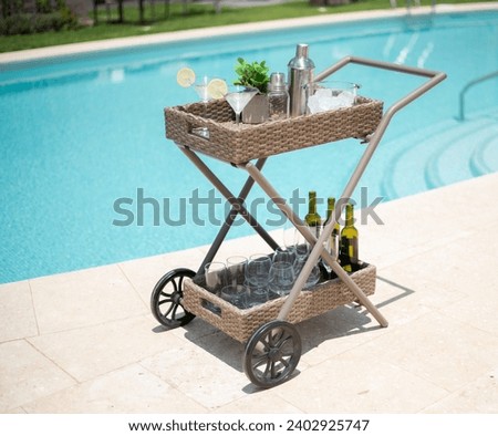 Summertime Poolside Celebration Cart Outfitted with a Rattan Bar Trolley Bearing Stemware, a Stainless-Steel Shaker and an Array of White Wine Bottles, Prepared for an Alfresco Party on Sunny Tiles. Royalty-Free Stock Photo #2402925747