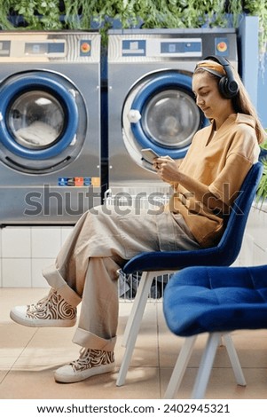 Young pretty woman in casualwear and headphones using smartphone while sitting on blue chair against automatic washing machines