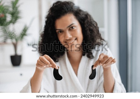 Focus on hands of charming multinational serene female with black eye gels wearing bathrobe and on home background. Lovely adult using anti-fatigue eye masks for skin hydration and glow.