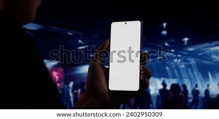 Caucasian woman holding a phone in two hands, people dancing in the night club in the background. Blank white screen smartphone mockup. Horizontal orientation