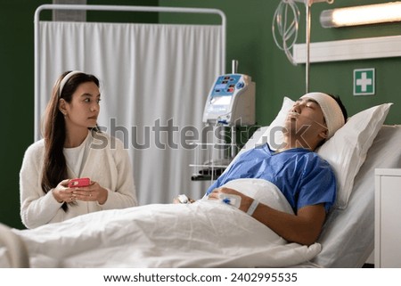 Next to her husband's bed in the hospital, the wife attentively watched, using her phone, providing support.