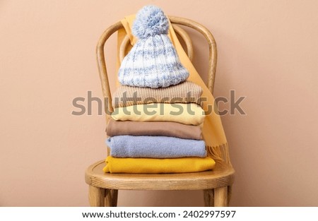 Stack of folded sweaters with hat and scarf on chair against beige background