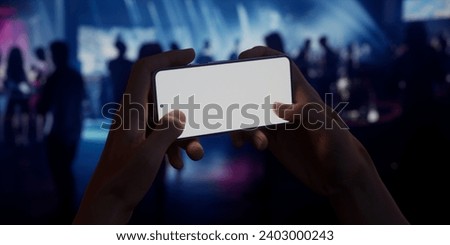 Caucasian man holding a phone in two hands, people dancing in the night club in the background. Blank white screen smartphone mockup. Horizontal orientation