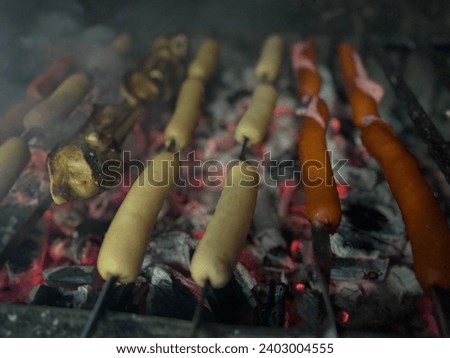 Grilled sausages on barbecue party night in backyard. Grill Pit With Glowing And Flaming Hot Charcoal Briquettes. Smoke from the fire. Tasty food smell. Grilled juicy sausages on a grill with fire.