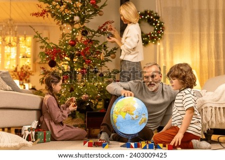dad and son play together on floor at home, examining globe, smiling and laughing, mom and daughter decorate Christmas tree on background
