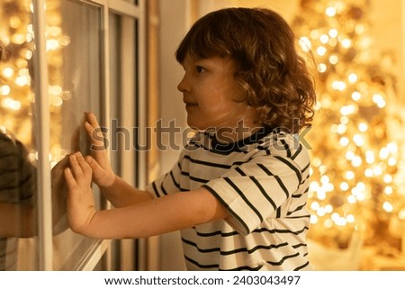 little boy with curly hair stand near window and look out, waiting santa claus with presents during christmas night
