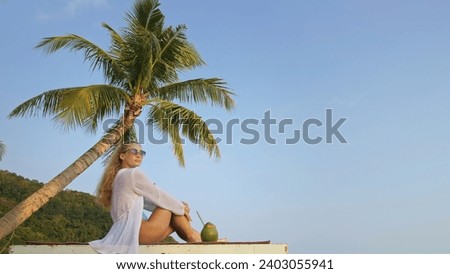Natural beauty woman is enjoy tropical view idyllic island. Woman spends her holidays on tropical island.Woman tourist with coconut cocktail on sunbed near palm tree.Concept trip exotic vacation rest