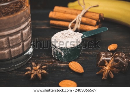 Whey protein powder in measuring spoon on dark wooden board with glass of milkshake smoothie drink, bananas, star anise, almonds, cinnamon stick, chocolate pieces.