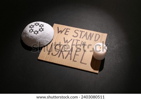 Text WE STAND WITH ISRAEL, Jewish hat and burning candle on dark background