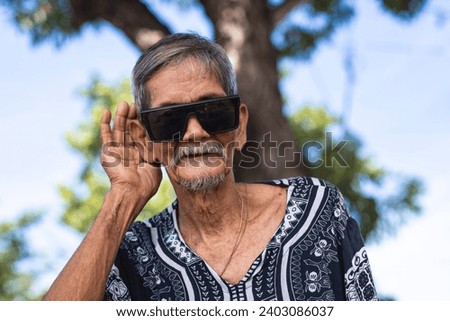 A cool hippie granddad tells someone to speak louder. Gesturing with his hands, possible deafness. Wearing shades and boho style shirt. Hanging out at the park. Royalty-Free Stock Photo #2403086037