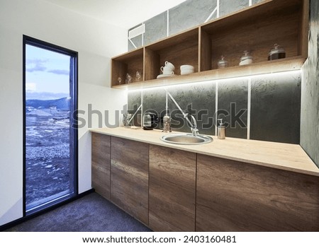 Modern kitchen with white walls and glass door with scenic view inside. Side view of kitchen area with wooden shelves and working surface, illuminated with warm LED light indoor. Concept of interior. Royalty-Free Stock Photo #2403160481