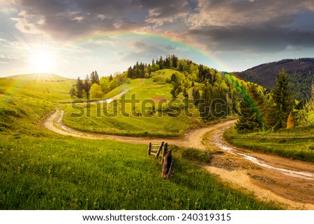 composite autumn landscape. fence near the cross road on hillside meadow in mountains. few fir trees of forest  on both sides of the road in sunset light with rainbow Royalty-Free Stock Photo #240319315
