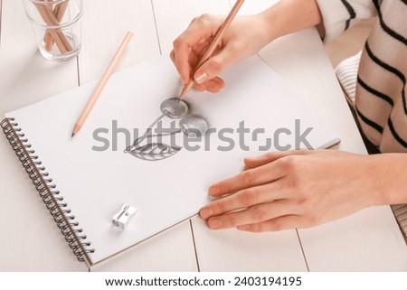 Woman drawing cherries with graphite pencil in sketchbook at white wooden table, closeup