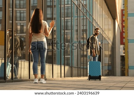 Long-distance relationship. Woman waving to her boyfriend with luggage near building outdoors Royalty-Free Stock Photo #2403196639