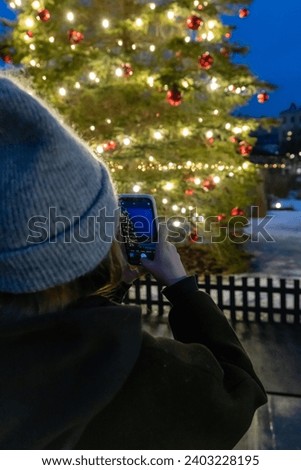 A girl takes a picture of a beautiful New Year tree on her phone in the evening. The Christmas tree is decorated with garlands and red balls in blur. Selective focus on phone