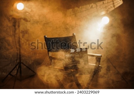 Close up shot of cinema clapperboard standing on director's chair, spotlights at the background, warm light and smoke in the scene. Royalty-Free Stock Photo #2403229069