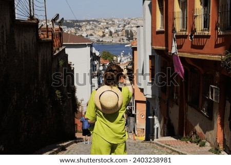 A girl tourist in a green summer suit and with a straw hat on her shoulders takes pictures of the cityscape in the center of the old part of Istanbul on her mobile phone. View from the back