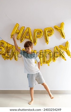 Boy in T-shirt, shorts, barefoot dancing against background of wall with Happy birthday inscription made of golden balloons.Layout of greeting card. Celebrating birthday. Positive emotions. 
